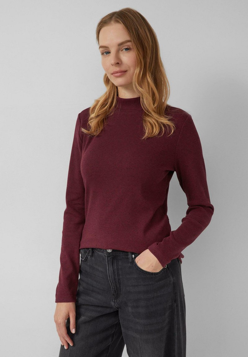 Vrouw met lichtbruin haar, gekleed in een bordeauxrode lange mouw top en zwarte jeans, staand met een hand in haar zak tegen een effen achtergrond.