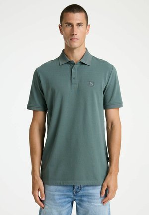 Man wearing a muted green short-sleeve polo shirt and light blue jeans, standing against a plain white background.