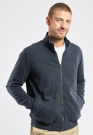 Homme aux cheveux courts portant une veste zippée bleu marine par-dessus une chemise blanche, debout les mains dans les poches devant un fond uni.