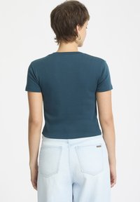 Türkise kurzärmelige Bluse aus Baumwolle, mit taillierter Passform und rundem Ausschnitt, kombiniert mit hellblauen, weit geschnittenen Jeans.