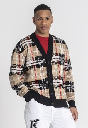 Cardigan a quadri beige con bordini neri, caratterizzato da dettagli rossi e bianchi, cinque bottoni e una vestibilità morbida, indossato sopra una maglietta rossa.