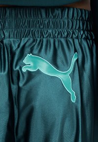 Κοντινή λήψη σπορ σορτς από γυαλιστερό ύφασμα σε τιρκουάζ χρώμα με ελαστική μέση και μεγάλο λογότυπο Puma σε ανοιχτό μπλε στο πλάι.
