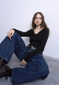 Γυναίκα με κυματιστά μαλλιά κάθεται στο πάτωμα φορώντας μαύρο μακρυμάνικο τοπ, παντελόνι широкий πόδι σε μπλε χρώμα, μαύρη ζώνη και σκούρα καφέ μπότες με ψηλό τακούνι.