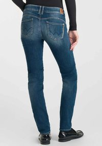 Persoon in aangesloten blauwe jeans en zwarte loafers, van achteren weergegeven met een zwart langgeraf shirt tegen een plain achtergrond.