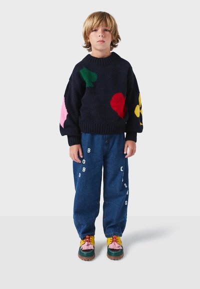 Marineblauer Strickpullover mit bunten abstrakten Formen, kombiniert mit blauen Jeans, die weißen Text tragen. Helle, mehrfarbige Schuhe runden das Outfit ab.