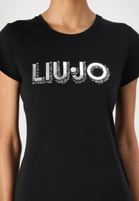 Musta T-paita lyhyillä hihoilla, jossa on hopeinen "LIUJO"-logo, koristeltuna pienillä kimalteilla ja sileällä pinnalla.