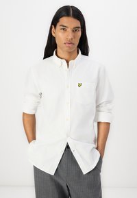 White button-up shirt made from a light fabric with a chest pocket and a yellow butterfly logo. Rolled sleeves and relaxed fit.