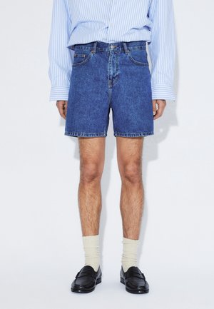 Pantalones cortos de mezclilla azul con un corte clásico, cinco bolsillos y costuras en contraste. Combinados con una camiseta de rayas de manga larga y mocasines.