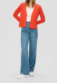 Röd cardigan över en vit skjorta, kombinerad med ljusblå vida jeans och vita platåskor. Tygerna är mjuka och avslappnade.