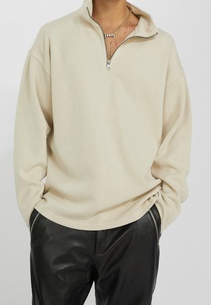 Persona con un jersey beige con cremallera hasta el pecho y mangas largas, y pantalones de cuero negros con bolsillos con cremallera, con las manos en los bolsillos.
