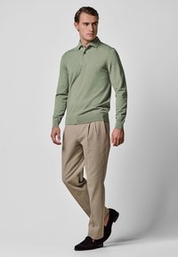 Hellgrünes Strick-Poloshirt mit klassischem Kragen, kombiniert mit beigen, plissierten Hosen und schwarzen Loafers, vor neutralem Hintergrund.
