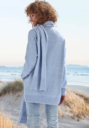 Hellblaue oversized Sweater mit geripptem Saum und Bündchen, drapiertem Schal, weicher Textur und lockerer Passform, getragen über hellen Jeans.
