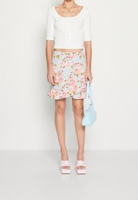 Žena v bílém krátkém zapínacím topu, modré květinové volánkové sukni, růžových platformových sandálech, držící světle modrou kabelku.