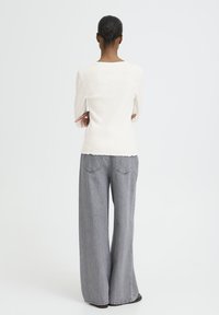 Sieviete ar bunā saviem matiem, ģērbusies baltā garo piedurknu krekliņā un pelēkos platās biksēs, stāv ar rokām saplākšņotas, vērsta prom no kameras.