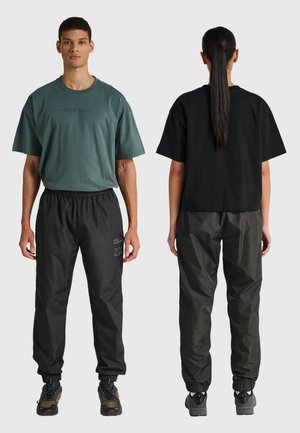 Twee mensen in casual kleding; één kijkt naar voren en draagt een groen t-shirt en een zwarte broek, de ander kijkt naar achteren en draagt een zwart t-shirt en een zwarte broek.