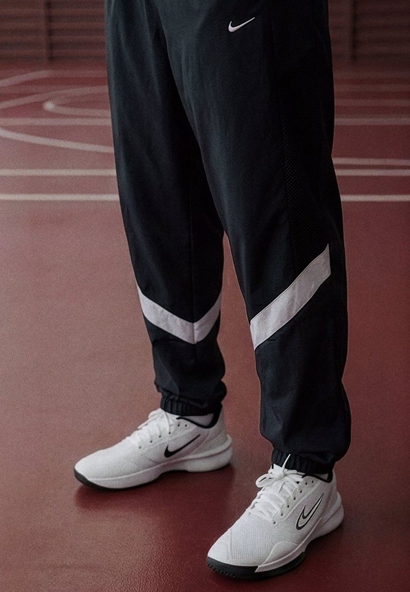 Jalat mustissa verkkareissa, joissa on valkoiset chevron-raidat, ja valkoisissa Nike-tennareissa punaisella sisäurheilukentällä.