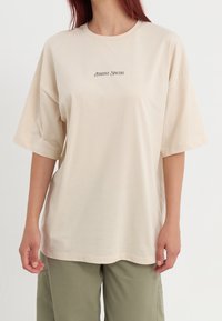 Camiseta oversized beige hecha de tela suave, con mangas cortas y escote redondo. El texto centrado dice "Positivo Sincero."