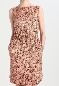Vestido sin mangas de durazno con manchas abstractas negras, cintura ceñida, bolsillos laterales y dobladillo a la altura de la rodilla, usado por una persona con las manos en los bolsillos.