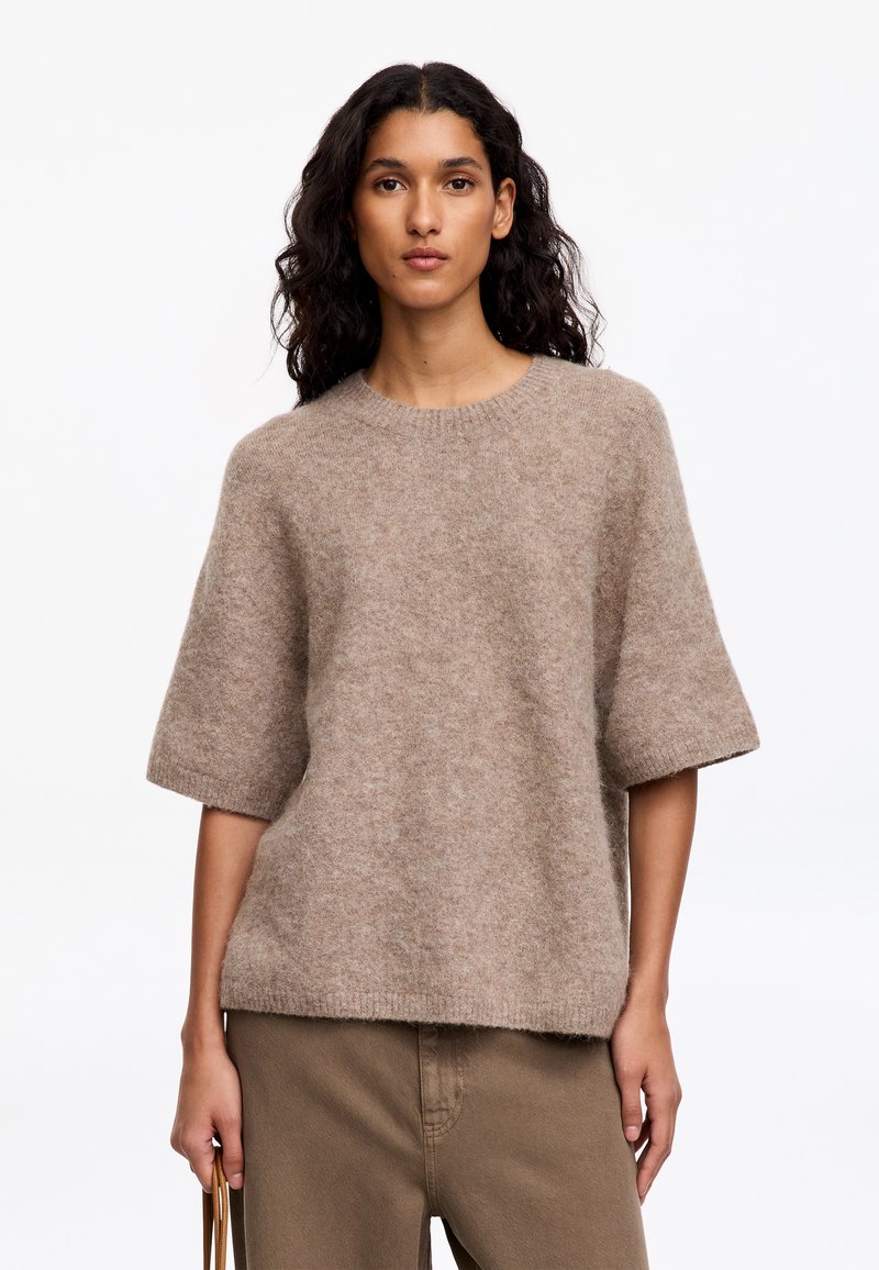 Femme aux longs cheveux bouclés foncés porte un pull beige ample à manches courtes et un pantalon marron, tenant une sangle de sac à main dans une main.