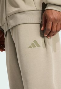 Beige sportkleding met een gestructureerde stof, voorzien van een opvallend olijfgroen Adidas-logo op het onderbeen. Close-up van ontwerpelementen.