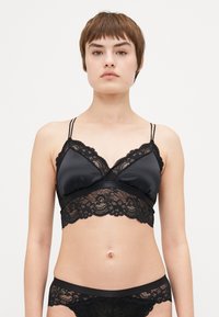 Svart bralette med sateng kropp og blondekant. Justerbare stropper, v-hals og bølget kant. Matchende svart blondeundertøy er synlig.