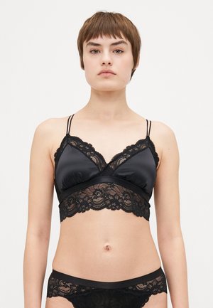 Black bralette with satin body and lace trim. Adjustable straps, v-neckline, and scalloped hem. Matching black lace underwear is visible.