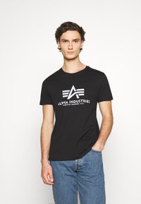 Sort bomulds T-shirt med rund halsudskæring, prydet med et hvidt logo og teksten "ALPHA INDUSTRIES" trykt på tværs af brystet. Stylet med blå jeans.