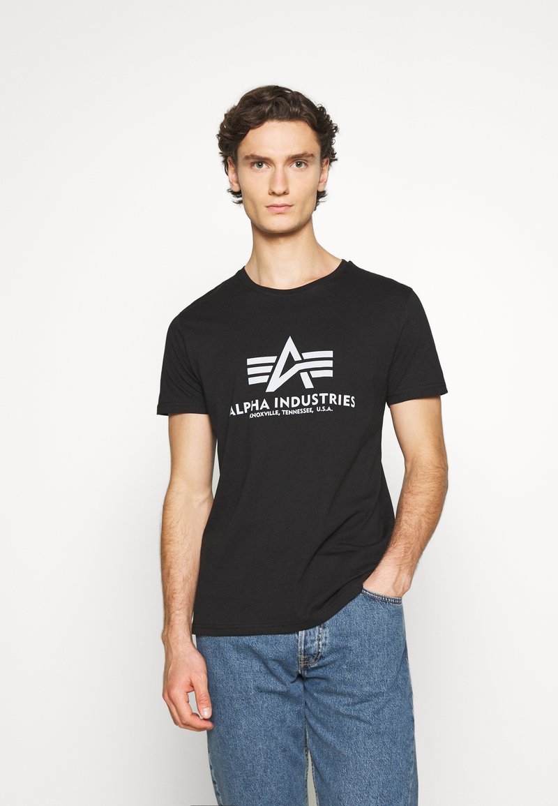 Sort bomulds T-shirt med rund halsudskæring, prydet med et hvidt logo og teksten "ALPHA INDUSTRIES" trykt på tværs af brystet. Stylet med blå jeans.