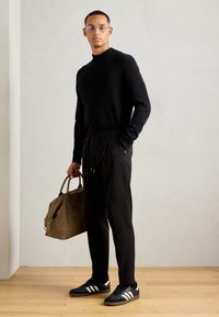 Man wearing black sweater, black pants, and black sneakers with white stripes, holding brown leather duffel bag, standing on wooden floor.