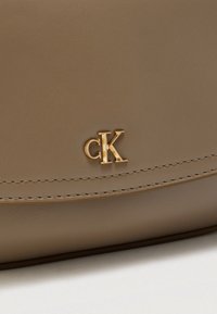 Beige Ledertasche mit glatter Textur, verziert mit einem goldenen "CK" Logo und sauberen Nahtdetails. Das Design ist minimalistisch und elegant.