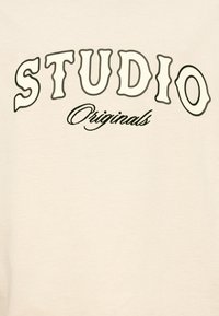 Ύφασμα κρεμ με τη λέξη «STUDIO» σε έντονα, κυματιστά γράμματα και το «Originals» με καλλιγραφική γραφή από κάτω. Διαθέτει λεία υφή και χαλαρή εφαρμογή.