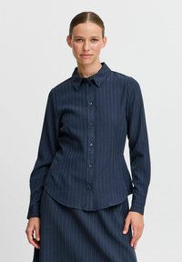 Camicia blu navy con bottoni, strisce bianche verticali, maniche lunghe, colletto a punta, orlo curvo e tessuto morbido.