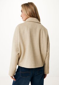 Beige kašmírová bunda s širokým límcem, uvolněným střihem a spadlými rameny. Má zkrácený design a viditelné švy na modrých džínách.