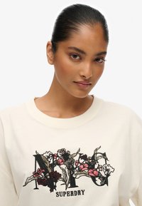 Felpa color crema con ricami floreali in tonalità di rosso e nero. Il design include la parola "SUPERDRY" sotto i fiori.