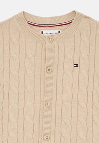 Béžový pletený kardigán so vzorom káblov, okrúhlym výstrihom a zapínaním na päť gombíkov. Obsahuje malý logový akcent.