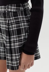 Bermudas de cuadro en blanco y negro con pliegues, que presentan una cinturilla estructurada, y una parte visible de una blusa negra de manga larga con canalé.