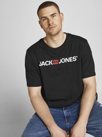 Musta puuvillapaita, jossa on valkoinen "JACK & JONES" -logo, yhdistettynä sinisiin farkkuihin. Paidassa on pyöreä pääntie ja lyhyet hihat.