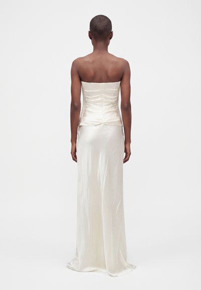 Personne aux cheveux courts portant une robe bustier en satin écru, debout de dos sur un fond blanc uni.