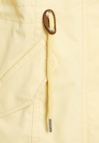 Close-up of pale yellow fabric with a braided drawstring, metal tip, leather patch, and stitched pocket detail.