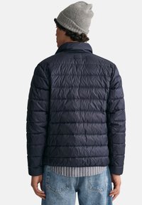 Mörkblå vadderad jacka med kviltad design och krage. Bärs över en grå- och vitrandig skjorta, tillsammans med ljusblå jeans.