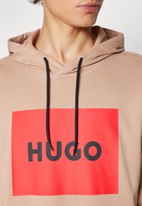 Sudadera con capucha de tela marrón claro. Presenta un gran gráfico rectangular rojo con "HUGO" en letras negras en negrita y cordones negros.
