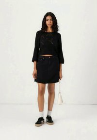 Mulher vestindo um suéter cropped preto, saia de ganga preta, meias brancas, tênis pretos, segurando uma pequena bolsa branca contra um fundo simples.