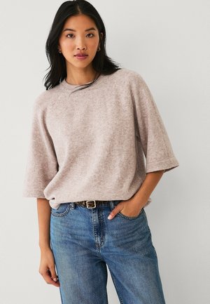 Ung kvinde iført en beige kortærmet sweater, stoppet ned i højttaljede blå jeans med et leopardmønstret bælte, stående foran en ensfarvet lys væg.