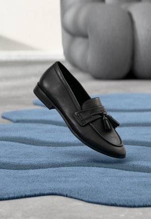 Black leather tassel loafer shoe floating above a textured blue rug with a gray upholstered chair in the background.