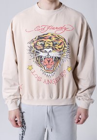 Sudadera beige con un gran gráfico de la cara de un tigre, con ojos verdes y lengua roja. Texto que dice "Ed Hardy" y "Los Ángeles."