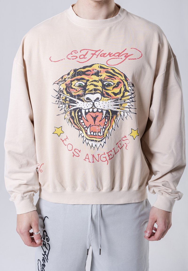 Sudadera beige con un gran gráfico de la cara de un tigre, con ojos verdes y lengua roja. Texto que dice "Ed Hardy" y "Los Ángeles."