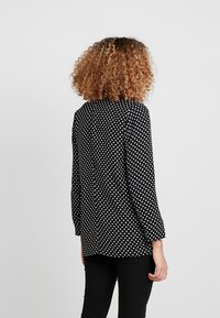 Czarna bluzka w białe kropki, z lekkiego materiału, z długimi rękawami, okrągłym dekoltem i luźnym krojem, widziana od tyłu.