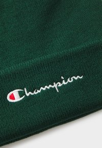 Zöld kötött sapka, fehér hímzett "Champion" logóval és egy kis piros-fehér akcentussal. Texturált anyag, hajtott széllel.