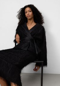 Kobieta z kręconymi włosami siedzi na czarnym krześle, ubrana w teksturowaną czarną sukienkę kopertową z satynowymi wykończeniami i paskiem, na tle białej ściany.