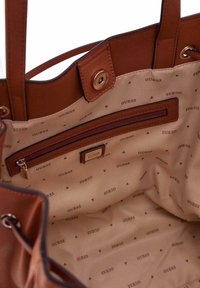 Braune Leder-Handtasche mit beigem Innenfutter, das wiederholt das "GUESS"-Logo und kleine dreieckige Muster zeigt; verfügt über ein Reißverschlussfach.